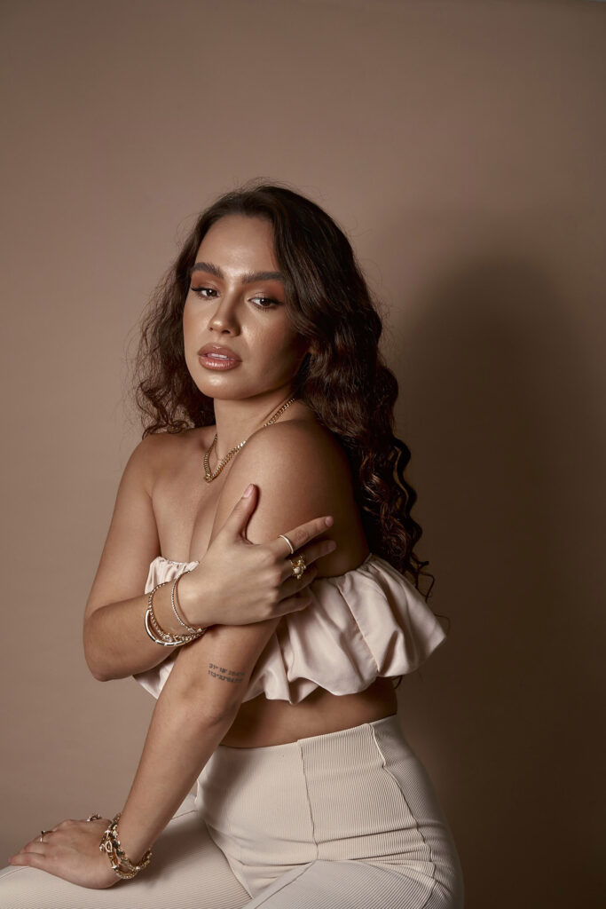 Abril Escalante a model with wavy brown hair is posed against a muted brown backdrop. She wears a white off-shoulder top and beige high-waisted pants accessorized with gold jewelry.